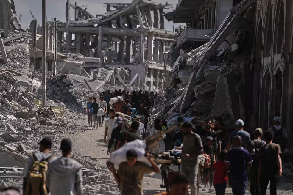 Palestinos deslocados caminham com seus pertences em frente a prédios destruídos enquanto retornam para suas casas em Khan Younis, no sul da Faixa de Gaza - Foto (AP Photo-Jehad Alshrafi)
